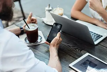 Two people sitting at a wooden table, one holding a smartphone, and the other using a laptop. A tablet lies on the table. They both have drinks; one is an iced coffee. The setting suggests a casual or work-related meeting in a cafe.