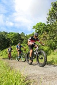 Three cyclists navigate their mountain bikes along a concrete path surrounded by greenery. The lead cyclist wears a black and red jersey, followed by two others in blue and black attire. The sky is partly cloudy, and the area is lush with trees and grass.