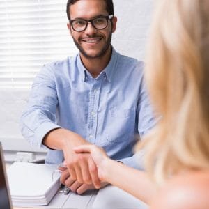 A man with glasses and a beard smiles while shaking hands with a woman across a desk, celebrating their new Career Transformation Package. The scene is an office with a laptop and documents.