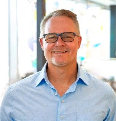 A person with short light hair and glasses is smiling indoors, exuding a coaching demeanor. They are wearing a light blue shirt with a collar. The background is blurred and bright, suggesting a well-lit room that sets the perfect framework for insightful conversations.