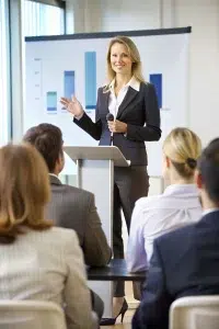A woman in a business suit stands at a podium, speaking to an audience in a conference room. A chart with bar graphs is displayed on a screen behind her as she discusses strategies to develop leadership potential.